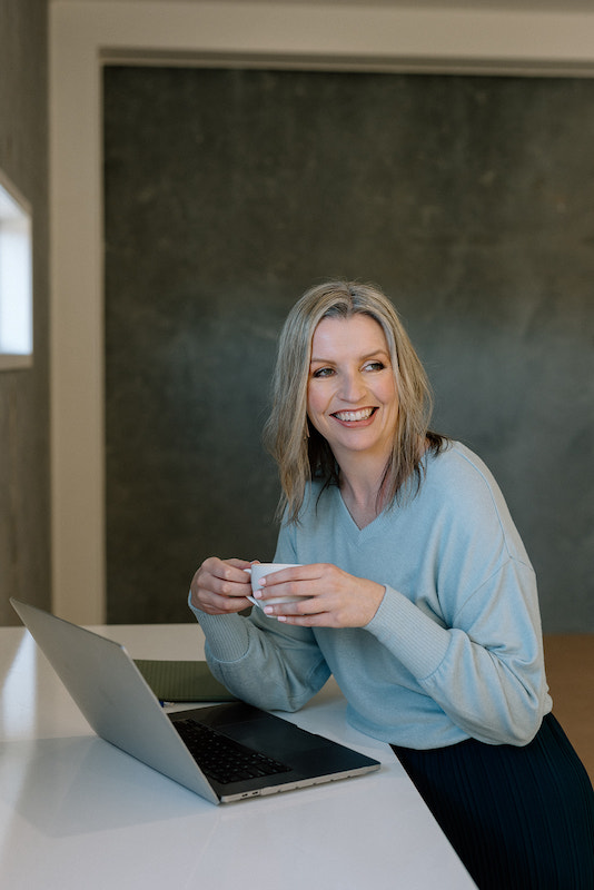 Donelle Dewar wearing a light blue long-sleeved top leaning against a table in front of a laptop. Donelle is holding a small cup of coffee.