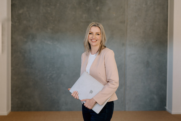 Image is of Donelle Dewar wearing dark pants, a white top and a light pink jacket. She's standing in front of a greenish-coloured wall holding an Apple laptop. She's an occupational therapist who helps employers with the benefits of employee wellbeing.