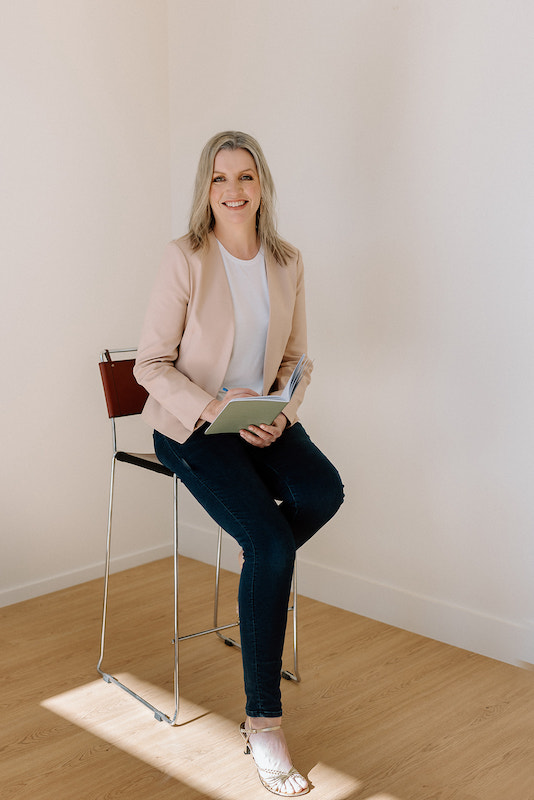 Image is of Donelle Dewar sitting on a high stool wearing dark jeans, a white shirt and a light pink jacket. She's holding a notebook in her left hand and a pen in her right hand. She's an occupational therapist who helps employees have good wellbeing at work.